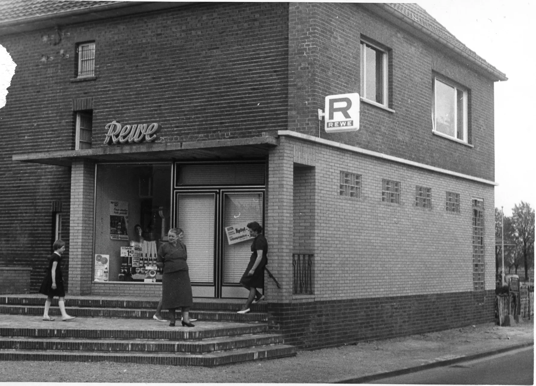 Historische Schwarz-Weiß-Aufnahme eines Ladenlokals mit Rewe-Schriftzug und Menschen auf der Eingangstreppe